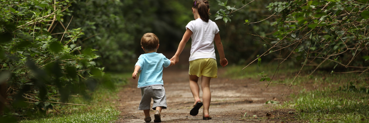 A brother and sister hold hands walking in a park